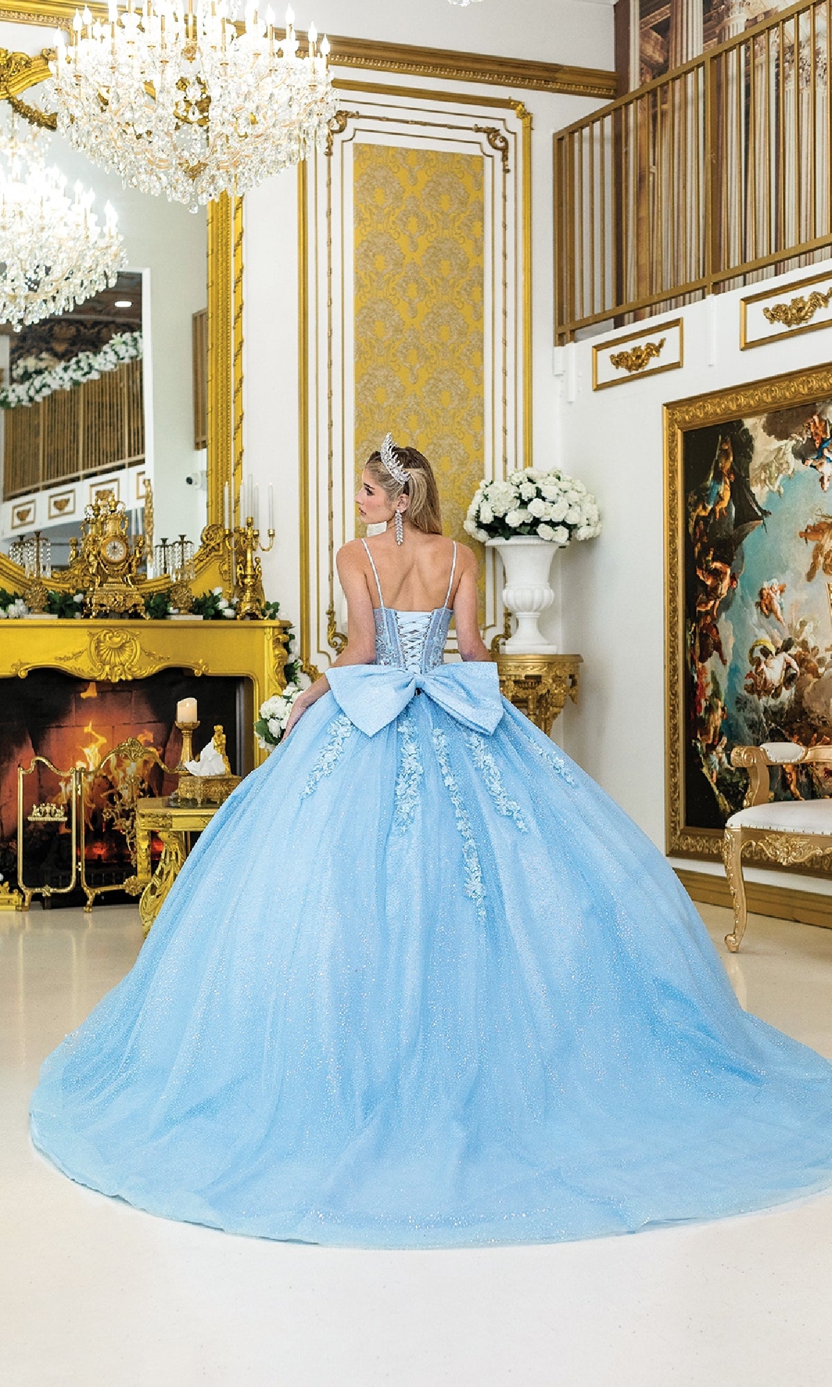 A woman in the Dancing Queen 1958 Long Quinceañera Dress with Bow stands facing a golden fireplace in an ornate room adorned with chandeliers and classic artwork.
